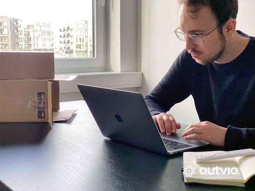 Zdjęcie przedstawia mężczyznę który sprzedaje online na platformie Amzaon ubrany jest w szary sweter siedzi przy laptopie a na biurku stoją przesyłki do wysłania w oknie widać bloki